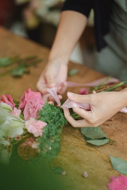 Close-up of a florist's hands arranging a bouquet of fresh flowers on a wooden table. The bouquet features pink roses, pale pink carnations, and lush green foliage, with some stems wrapped in pink flo