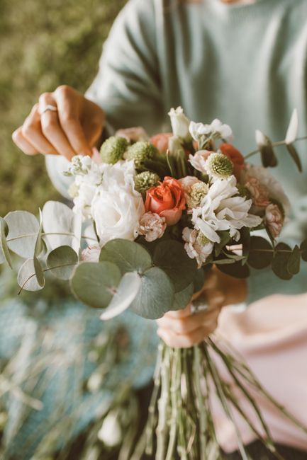 A person holding a mixed floral bouquet composed of white, peach, and light pink blossoms, including roses, lisianthus, and chrysanthemums, accented with soft green eucalyptus leaves. The bouquet is l