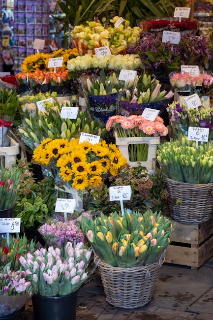 An array of fresh flowers displayed at a florist's market stall, featuring bright yellow sunflowers, pink tulips, purple tulips, white freesias, and pale pink daisies arranged in various baskets and p