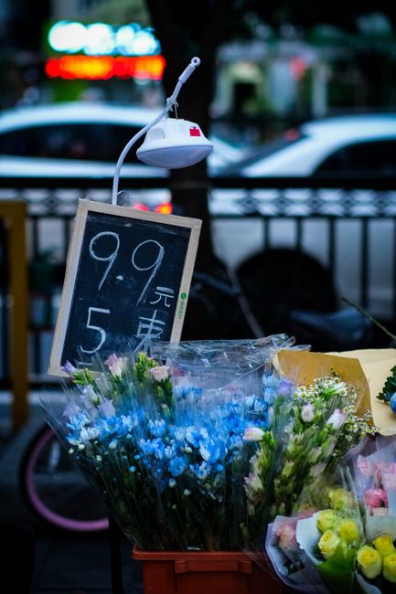 A display of fresh flower bouquets arranged outdoors on a stand, featuring a variety of flowers including blue and white blossoms, with some yellow flowers visible. The flowers are wrapped in clear pl