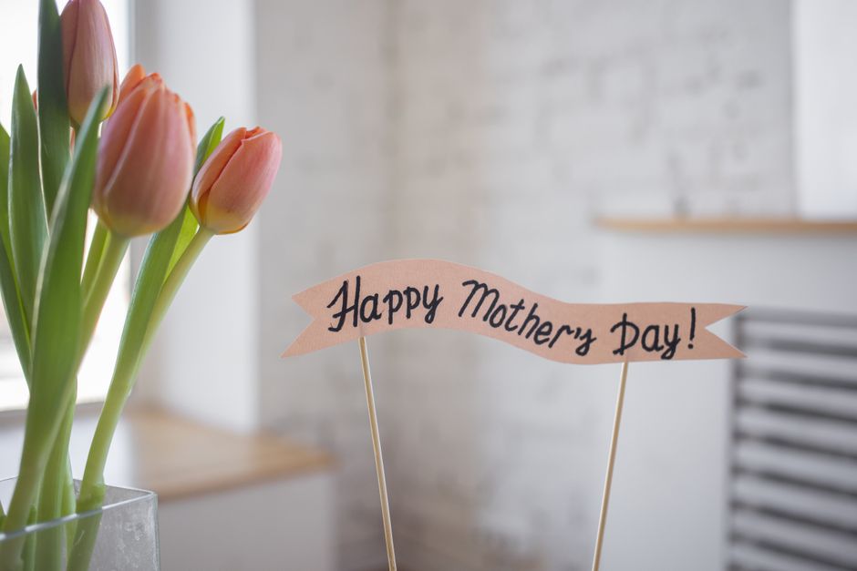 A bouquet of fresh pink tulips with green stems and leaves, arranged in a clear glass vase placed on a wooden surface. The tulips are in full bloom, showcasing smooth, rounded petals. Next to the bouq