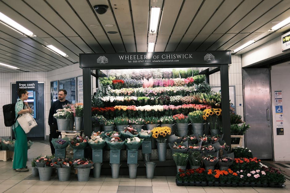 A vibrant floral display at Wheeler of Chiswick in an underground setting, featuring a variety of fresh flowers arranged in tiers, predominantly in shades of white, pink, red, yellow, and purple. The