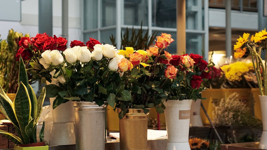 A collection of fresh flower arrangements displayed on a surface, including a bouquet of red roses and white lisianthus in a metallic silver vase, alongside yellow and orange roses in a cream-colored 