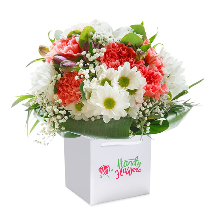 This radiant bouquet offers a true taste of Wapping's floristry craftsmanship, strikingly presented in a neat, modern white gift bag with soft ribbon handles-an ensemble ready to brighten any occasion from Canal Bridge to Tobacco Dock. The rounded arrangement brims with plush coral-pink and vibrant red carnations whose signature ruffled petals create a rich, inviting texture. Bright white daisies take pride of place among the carnations; their large, snowy petals and lively lime-hued centers radiate optimism and bring the freshness of a springtime walk along Wapping Wood. Wisps of delicate white baby's breath float between the heartier blooms, infusing the bouquet with a gentle, cloudlike lightness that catches the soft glow of daylight. Interspersed among the open flowers, a few gently closed buds-dusted with hints of purple and magenta-add a layer of intrigue and dimension. Encircling the composition, deep green glossy leaves and abundant foliage shape the bouquet into a full, rounded silhouette, grounding the vibrant colours and bestowing an elegant, harmonious finish. The immaculate white background and chic gift bag heighten the flowers' natural splendour, making this arrangement an ideal choice for birthdays, anniversaries, or uplifting gestures delivered seamlessly by Wapping's passionate local florists. Every petal and leaf is thoughtfully placed to evoke a sense of freshness, abundance, and heartfelt intention, perfectly suited for marking memorable moments in the heart of Wapping.