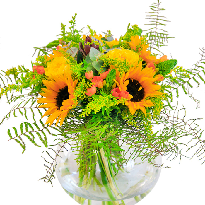 The first impression of this abundant bouquet is one of radiant, sunlit exuberance-a fitting tribute to the lively spirit of Wapping itself. Large, golden sunflowers spread open with dazzling intensity, their textured brown centers anchoring the bouquet much as the local warehouses anchor the riverside. Flanking them, deeply layered yellow garden roses burst into bloom, their petals offering a tactile softness that invites closer appreciation, like the gardens hidden down the cobbled local avenues. Tucked between the blooms are glossy scarlet Hypericum berries, sparkling against the lush, textured greenery that spills out in gentle, cascading fronds. Wisps of fern add feathery movement, while delicate fillers-perhaps goldenrod-introduce clouds of tiny, fresh yellow blossoms and slender green spikes, reminiscent of the wild plants seen along the canal paths. Blue-green eucalyptus leaves peek through, their matte finish a graceful complement to the arrangement's vivid palette. The round glass vase, perfectly transparent, frames the green stems immersed in water, highlighting the bouquet's structured grace and the natural beauty within. The overall presentation is bright and hopeful, carrying with it the scents and textures of summer open spaces found around Wapping. Ideal for celebratory occasions-birthdays, anniversaries, or simply brightening a beloved home-this bouquet brings a sense of happiness and local character into every space it graces, from riverside flats to homes overlooking Tobacco Dock.