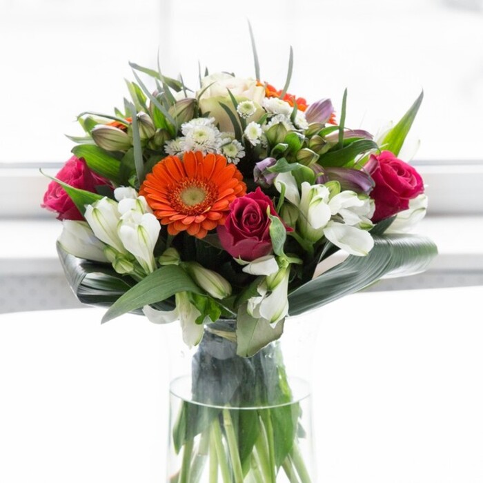 Carefully arranged to highlight both structure and emotion, this vivid bouquet entwines the richness of orange and peach gerberas with the luxurious magenta of freshly cut roses, all bathed in gentle, natural light reminiscent of Wapping's riverside apartments on a clear day. The orange gerberas burst forth with energy, their petals fanning out from dark central discs, while creamy, soft peach gerberas introduce a subtler glow, balancing the palette. The deep magenta roses, hints of their sweet scent mingling with the air, nestle gracefully among bright white alstroemeria and petite filler blooms. Each layer of the arrangement unveils new textures-the broad, glossy aspidistra leaves enveloping the lower half suggest a nurturing embrace, reminiscent of Wapping's close-knit community. Delicate sprigs of fresh greenery weave throughout, supporting and enhancing the colorful display. Clustered within a tall, clear glass vase, the vivid stems draw the eye downward, their clean lines and visible structure underscoring the arrangement's freshness and elegance. Light diffuses through the bouquet, capturing subtle shadows and enhancing the vibrancy of every petal. This ensemble would sit perfectly on a counter in a Wapping home or café, infusing the space with a spirit of celebration, gratitude, and warmth-ideal for new beginnings, expressing appreciation, or sending thoughtful wishes for happiness.