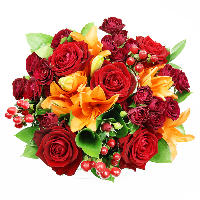 Seen from above on a stark white surface, this lush, circular bouquet reads like a curated study in reds and oranges, hand-tied to maximize visual impact and tactile delight. The composition is anchored by several large, fully bloomed deep red roses - each with plush, velvety petals that fold inward toward shadowed centres, evoking classic romance and a luxurious softness. Bright orange Asiatic lilies punctuate the arrangement with trumpet silhouettes and a sunny intensity; some lilies open wide to reveal their stamens and subtle speckling, while others remain in graciously curved buds, offering a sense of unfolding narrative. Small clusters of deep burgundy spray roses provide concentrated, velveteen texture and a darker counterpoint that enriches the colour story. Scattered among the flowers are glossy hypericum berries in a vivid ruby tone, their round surfaces reflecting the light and adding a lively, tactile accent. Broad green foliage - likely ruscus or salal - peeks out between blooms, cooling the palette and enhancing the bouquet's three-dimensional fullness. The soft, even lighting preserves delicate textures: the satin sheen of rose petals, the firm gloss of berries, and the subtle grain of foliage. A florist arranging in Wapping Lane or near the Prospect of Whitby pub might choose this palette to celebrate an autumn wedding, a heartfelt anniversary, or to lift a flat on the river with something richly joyful and inherently local; the bouquet speaks of warmth, celebration, and the quiet craftsmanship of neighbourhood floristry.